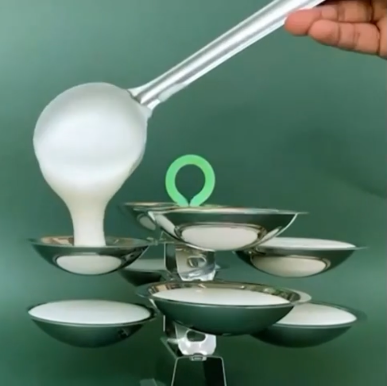 A hand holds a metal ladle pouring idli batter into an idli tray.